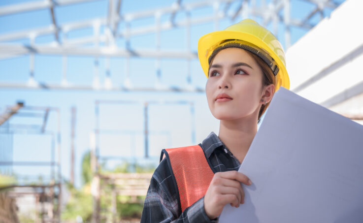 Asian female engineer wear yellow safety hard hat standing and holding blueprint while checking infrastructure 