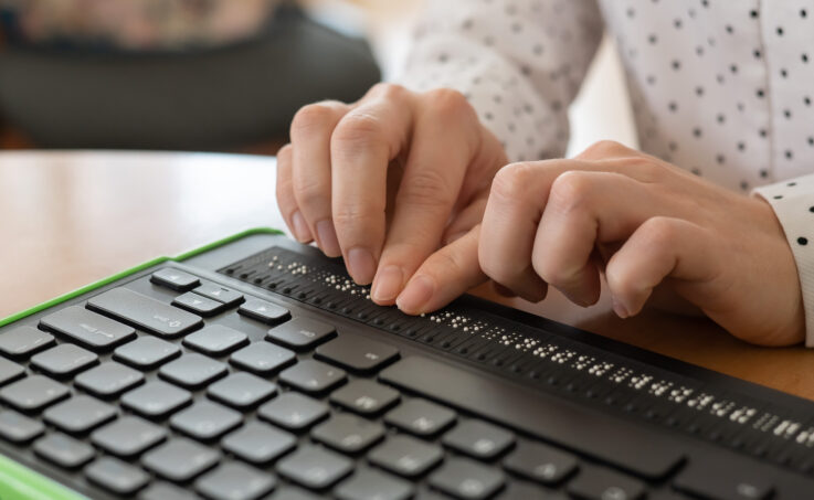 A blind woman uses a computer with a Braille display and a computer keyboard. Inclusive device.