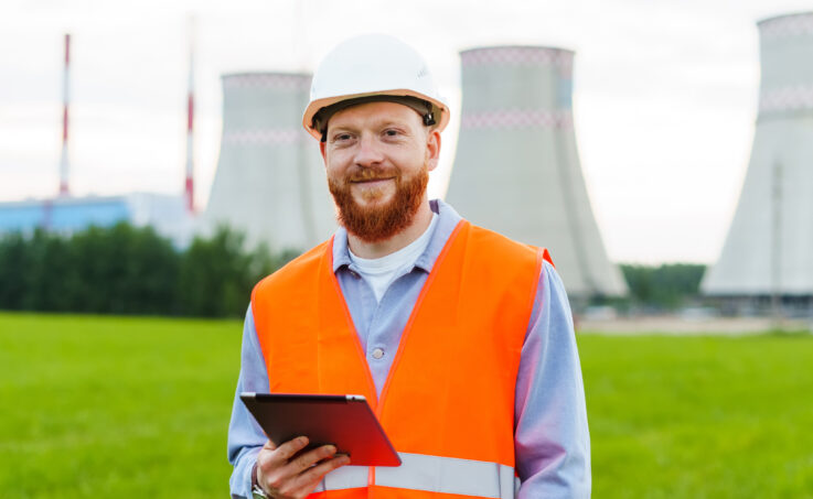 Nuclear power plant engineer with a tablet, standing in front of the cooling towers of a nuclear power plant