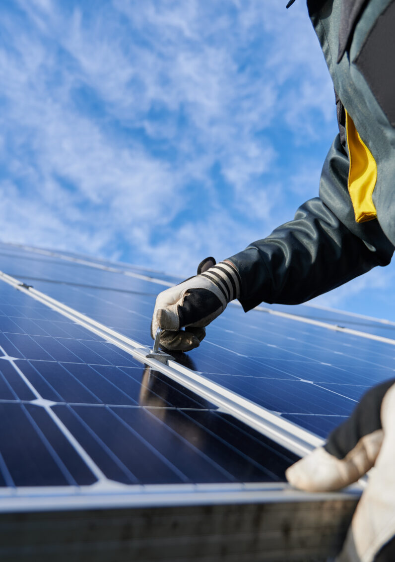 Close-up of technician with work gloves installing stand-alone photovoltaic solar panel under blue sky with clouds.