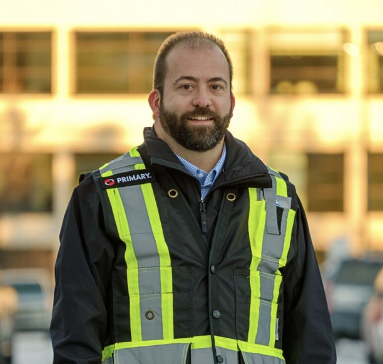 A man with a beard wearing a high-visibility vest stands outdoors in front of a building, smiling at the camera.