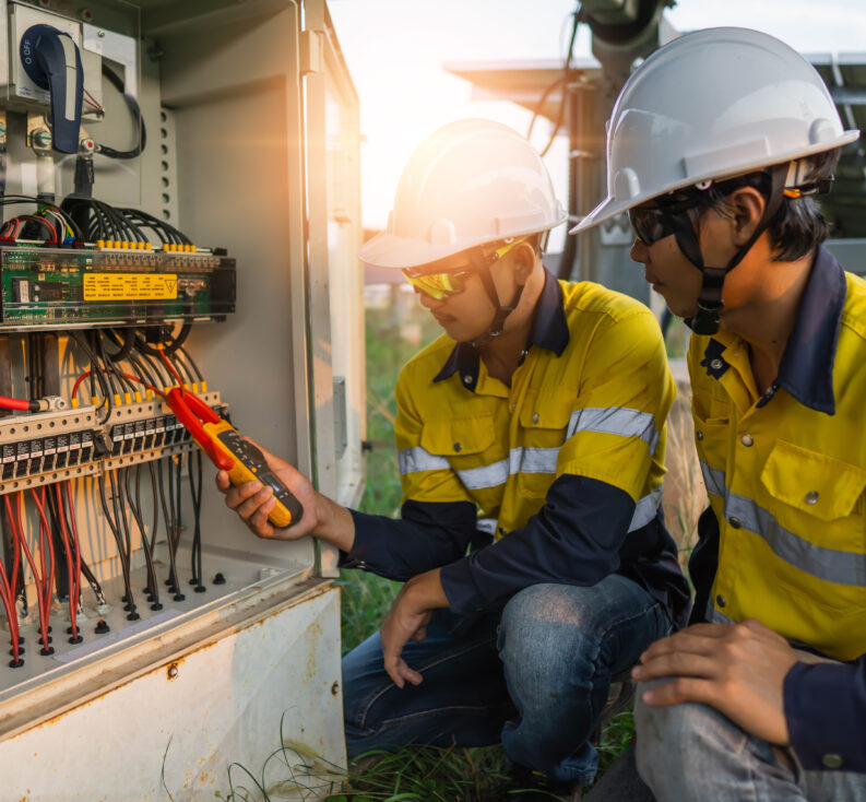 Two electricians in safety vests, hard hards and glasses looking at an electronic reader