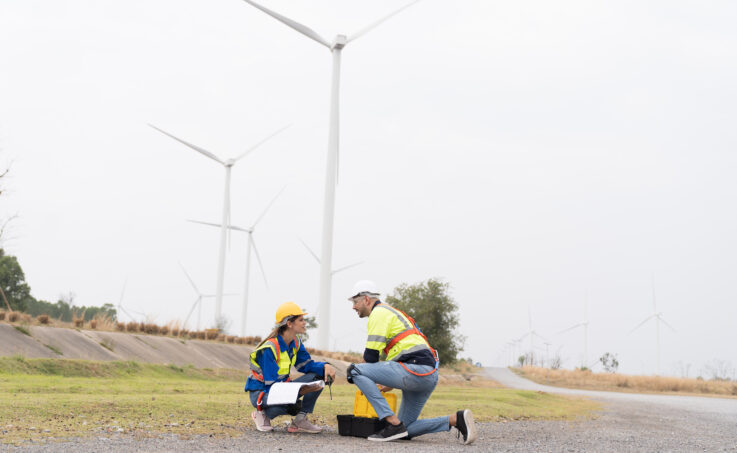 Two workers kneel outside with a toolbox and take notes, with multiple wind turbines in the background