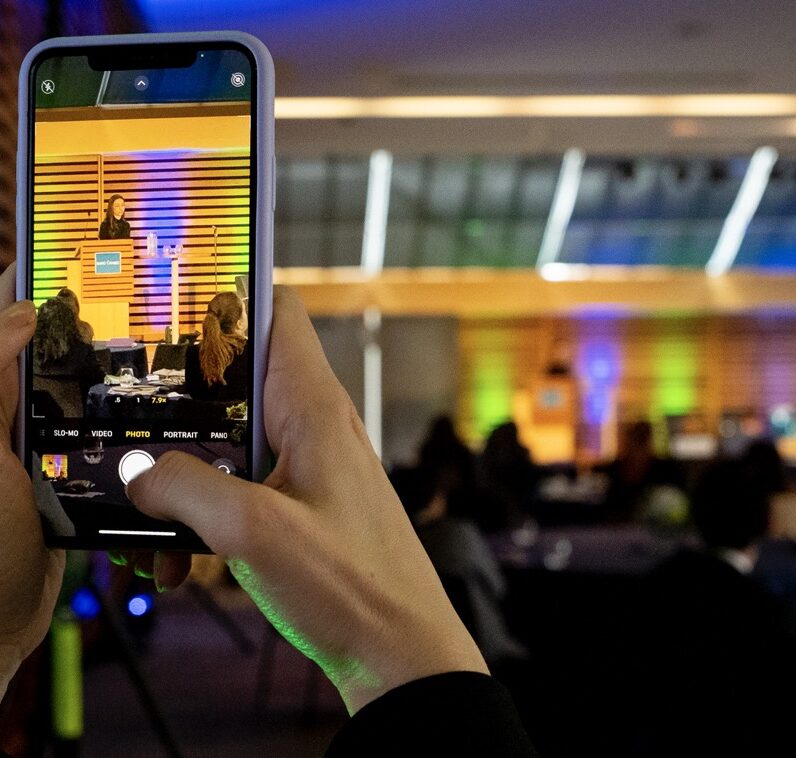 A person takes a photo with their smartphone of the presenter at the podium