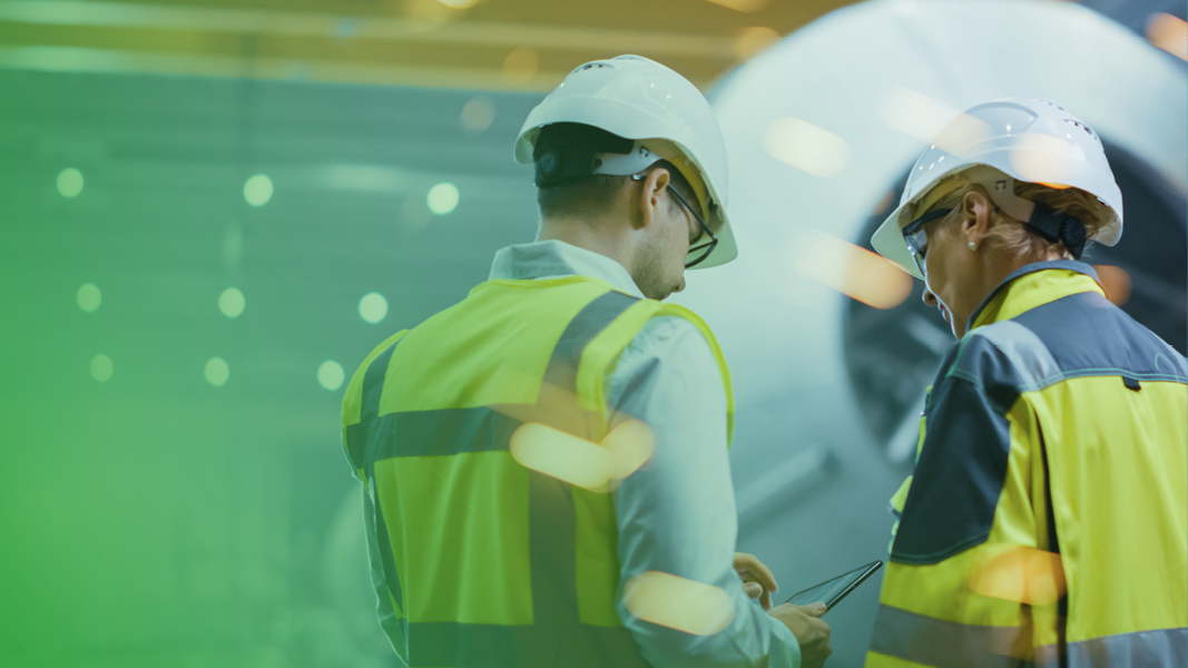 Two workers in safety vests and helmets discuss something while looking at a tablet in an industrial setting.