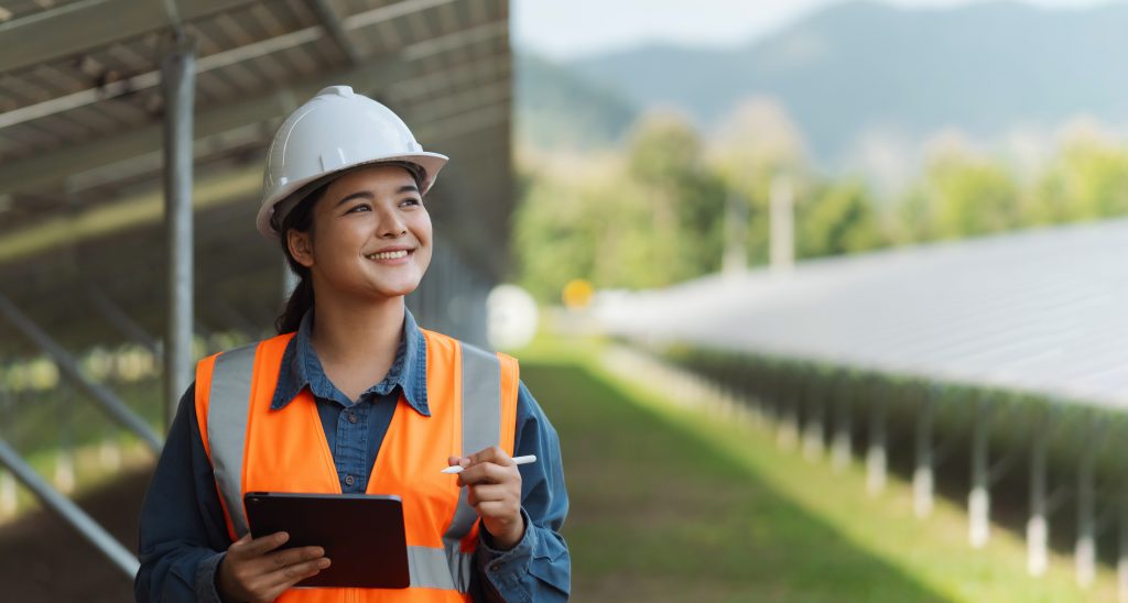 Person wearing a hard hat and orange safety vest holding a tablet at a solar energy facility.