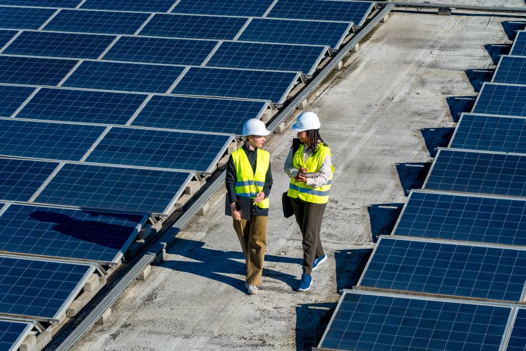 Two workers in safety vests and hard hats walk between rows of solar panels.