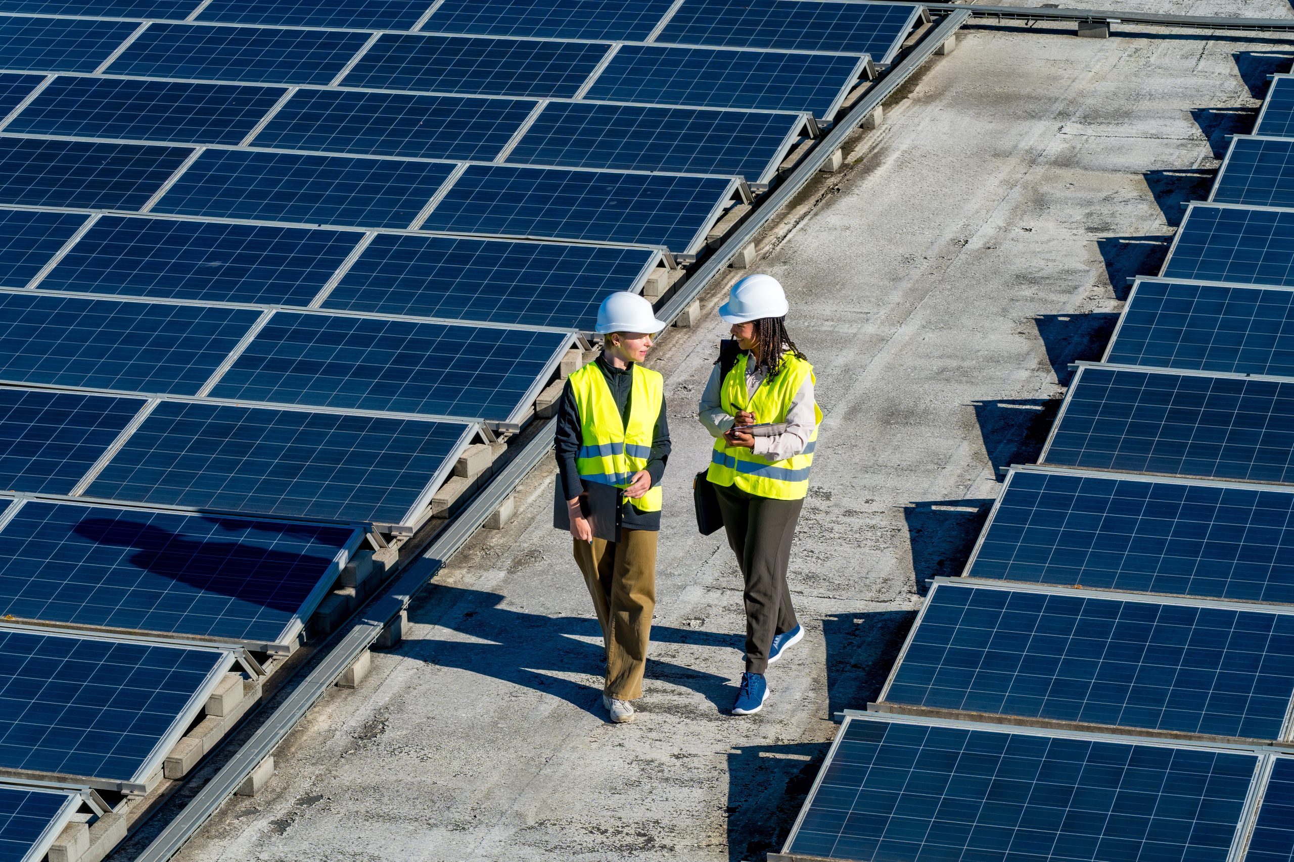 Two workers in safety vests and hard hats walk between rows of solar panels.