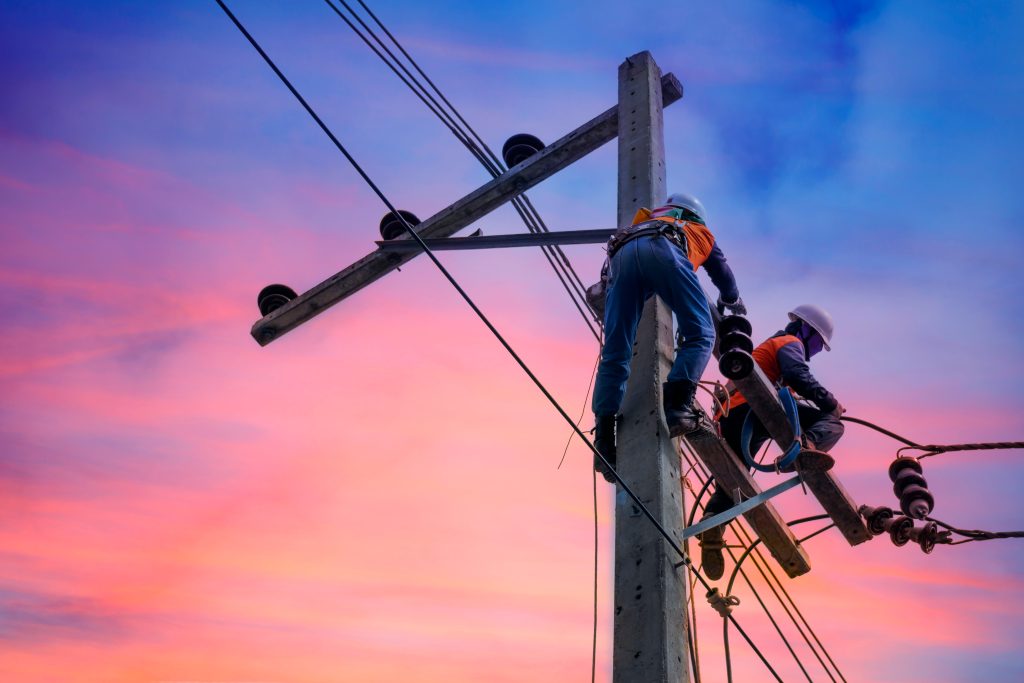 Lineworkers in safety gear climb and work on a pole with overhead wires, set against a colorful sunset sky.