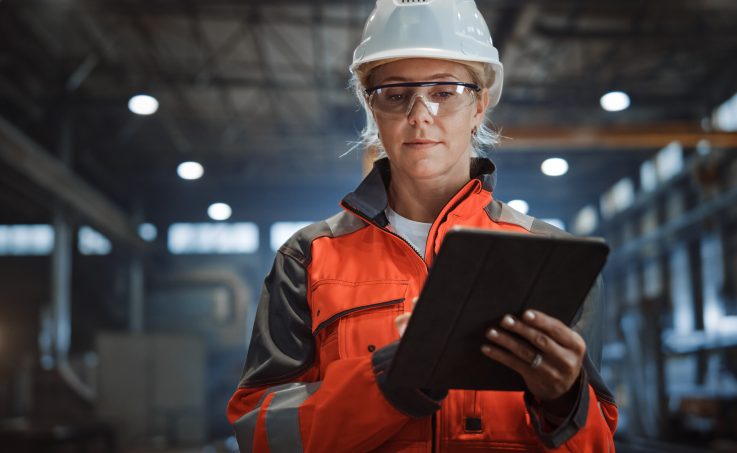 Worker in safety gear using a tablet in an industrial facility.