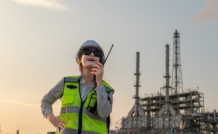 Worker in hard hat and safety vest using a two-way radio at an industrial facility.